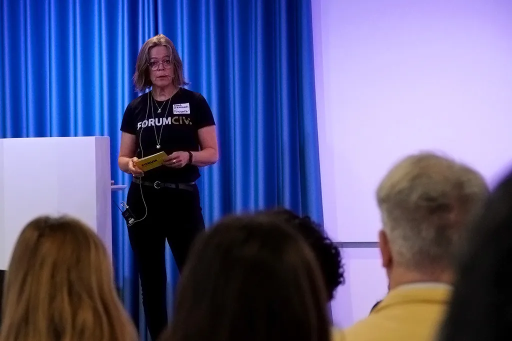 Anna talar till publiken på en scen. Hon har en svart t-shirt med texten ForumCiv.